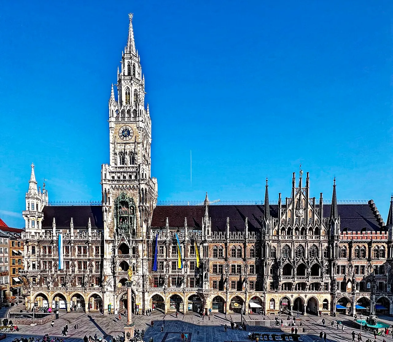 A torre gótica do Neues Rathaus na Marienplatz, com relógio e carrilhão, bandeiras penduradas na fachada e gente na praça, sob céu azul sem nuvens.