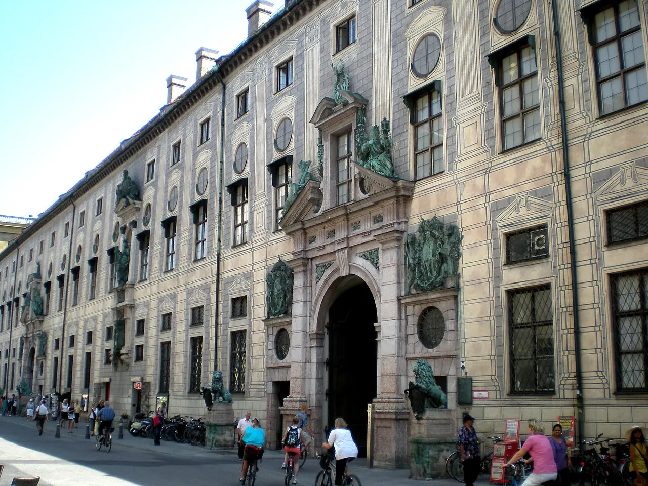 Fachada ornamentada da Residenz de Munique, com portal em arco guardado por estátuas de leões de bronze, esculturas na sacada e bicicletas estacionadas na rua.
