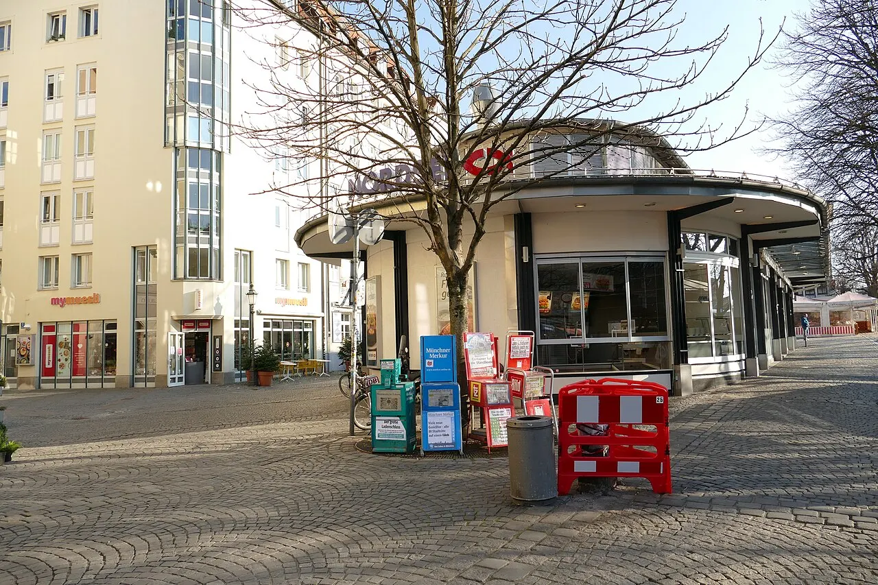 Quiosque de telhado redondo no Viktualienmarkt, em Munique, com caixotes e bancas de jornal na frente e a praça vazia.