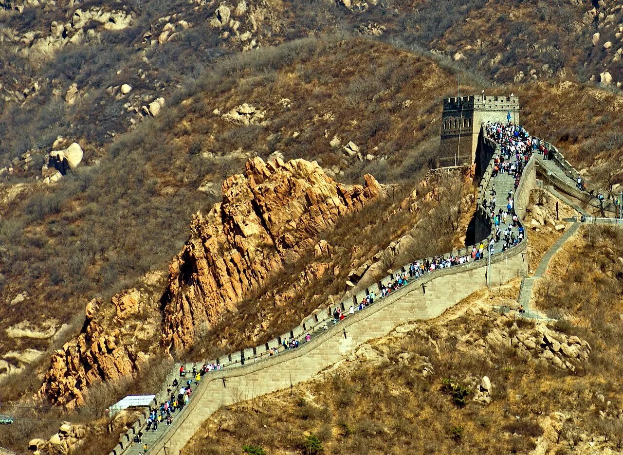 Vista aérea de um trecho da Muralha da China subindo a montanha até uma torre de vigia, com multidão de visitantes caminhando pelo caminho de pedra.