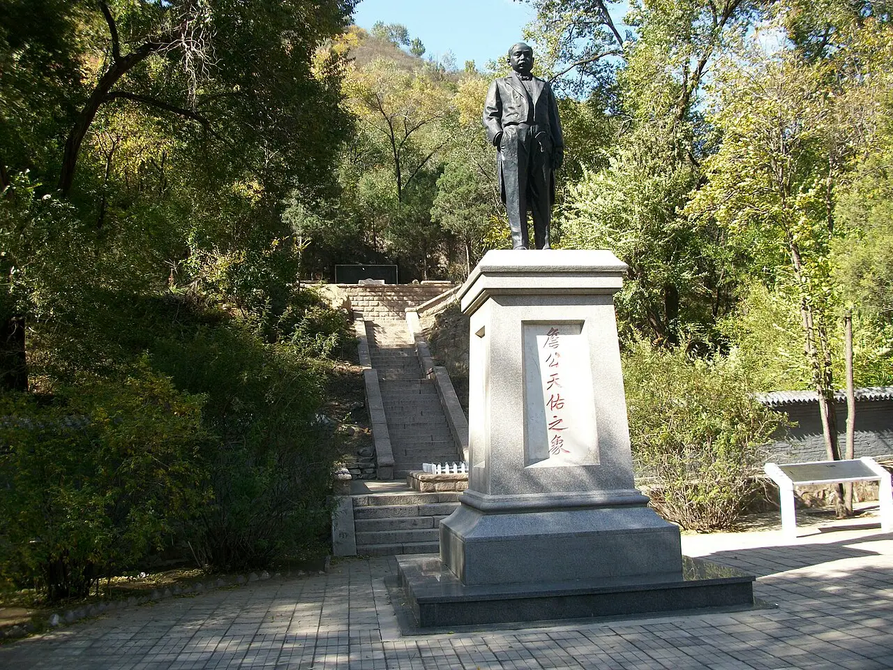 Estátua de homem em pé sobre um pedestal de pedra com inscrição vermelha, cercada de árvores.