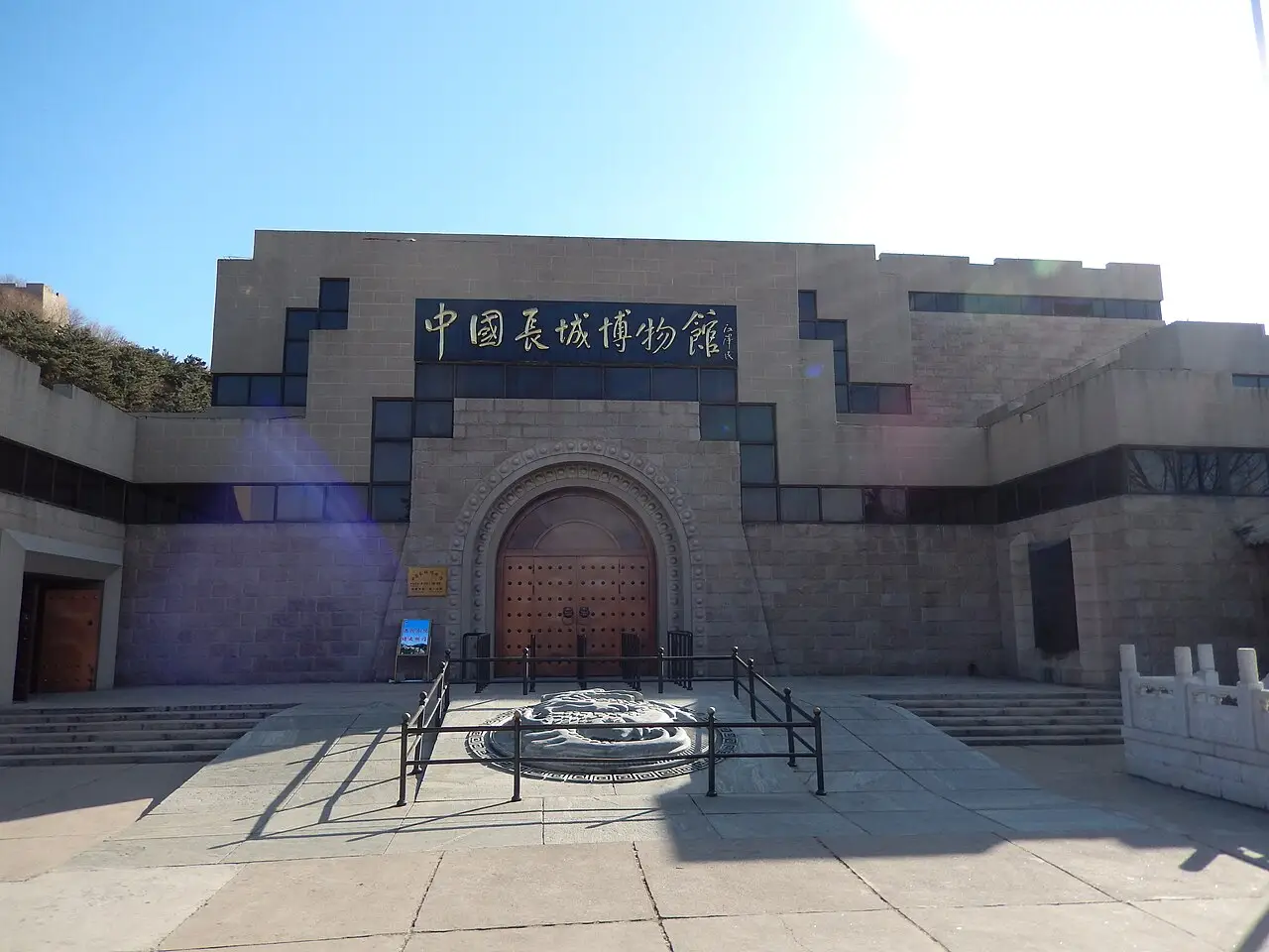 Fachada do Museu da Grande Muralha, de pedra clara, com o nome em chinês sobre o arco e um pátio de lajes na frente.