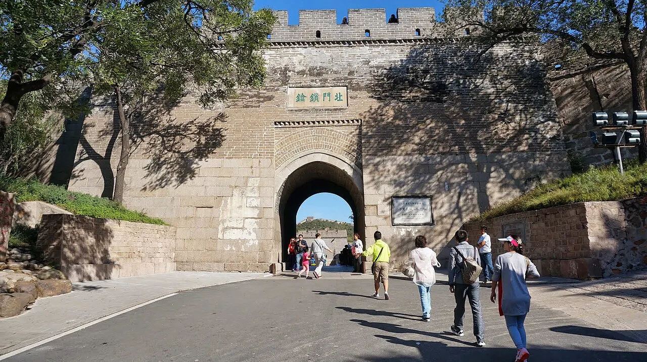 Portão de pedra com arco e ameias no alto, parcialmente sob galhos de árvore, com pessoas passando por baixo.