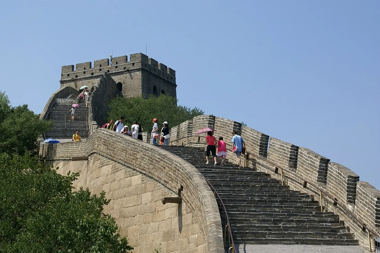 Trecho íngreme da Muralha da China com degraus de pedra subindo até uma torre de vigia, visitantes caminhando com guarda-sóis sob céu azul.