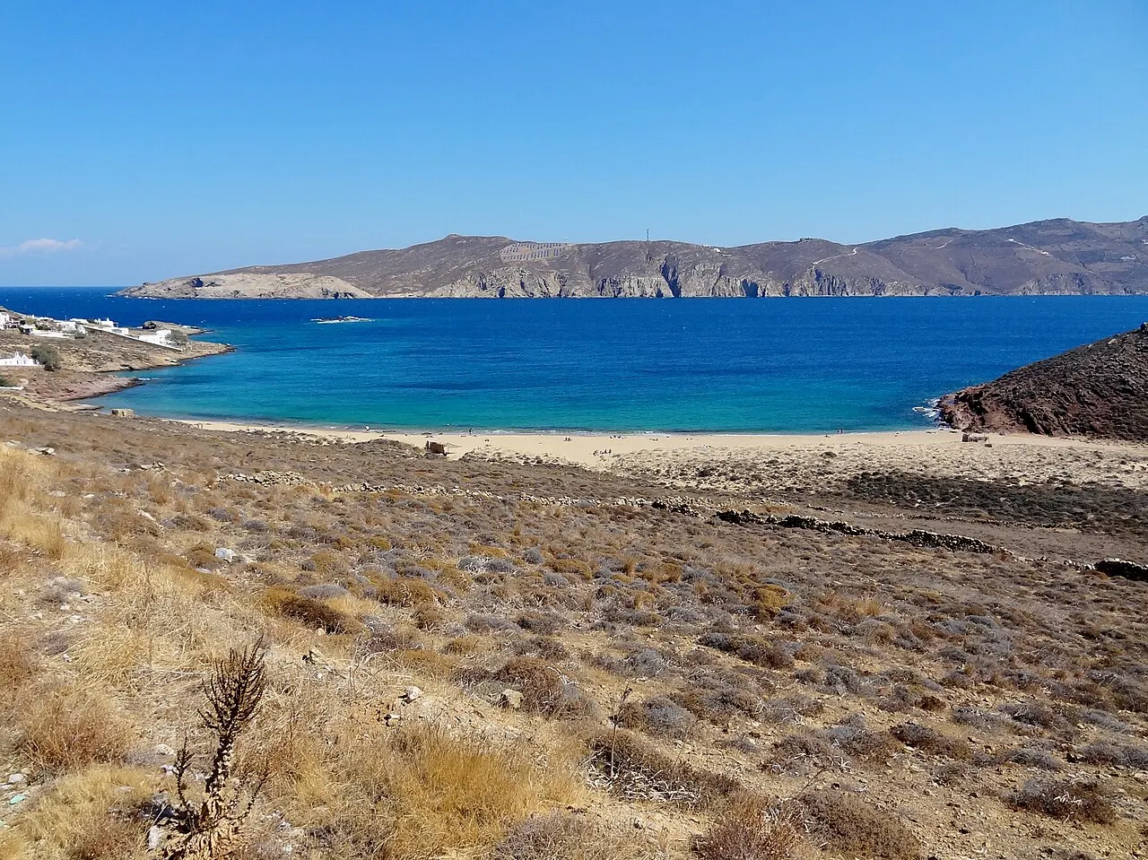 A baía de Agios Sostis, na costa norte de Mykonos, com areia clara, mar azul-escuro e encostas secas quase sem construções, sob céu azul.
