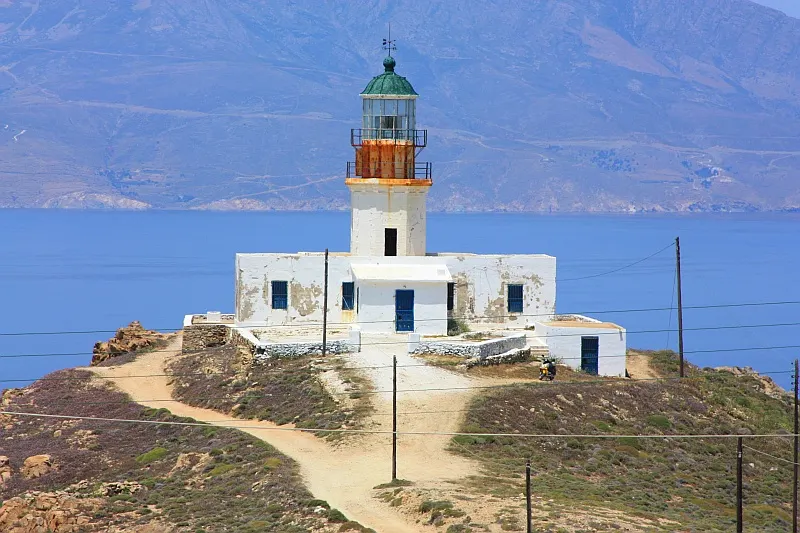 O farol de Armenistis, uma torre branca com lanterna verde sobre uma casa baixa, no alto de um promontório seco, com o mar azul e a ilha de Tinos ao fundo.