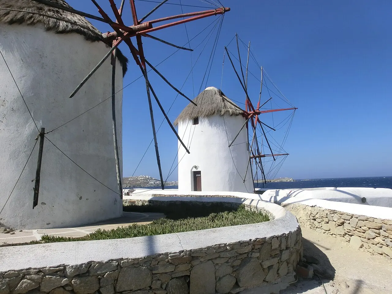 Dois moinhos de vento brancos e redondos de Kato Mili, com telhado de palha e pás de madeira, sobre um muro baixo de pedra, com o mar azul ao fundo.