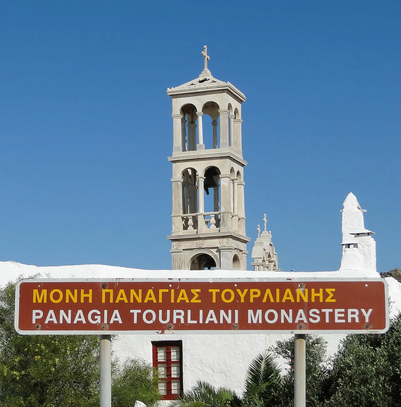 Torre de pedra clara do mosteiro de Panagia Tourliani, com arcos, sinos e cruz no alto, atrás de um muro branco e de uma placa vermelha com o nome do mosteiro em grego e inglês.