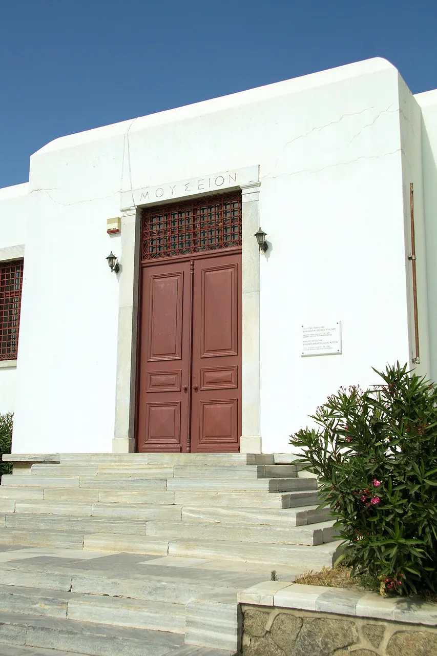 A entrada do Museu Arqueológico de Mykonos, um prédio branco com porta de madeira avermelhada no alto de uma escada de pedra, com um arbusto ao lado.