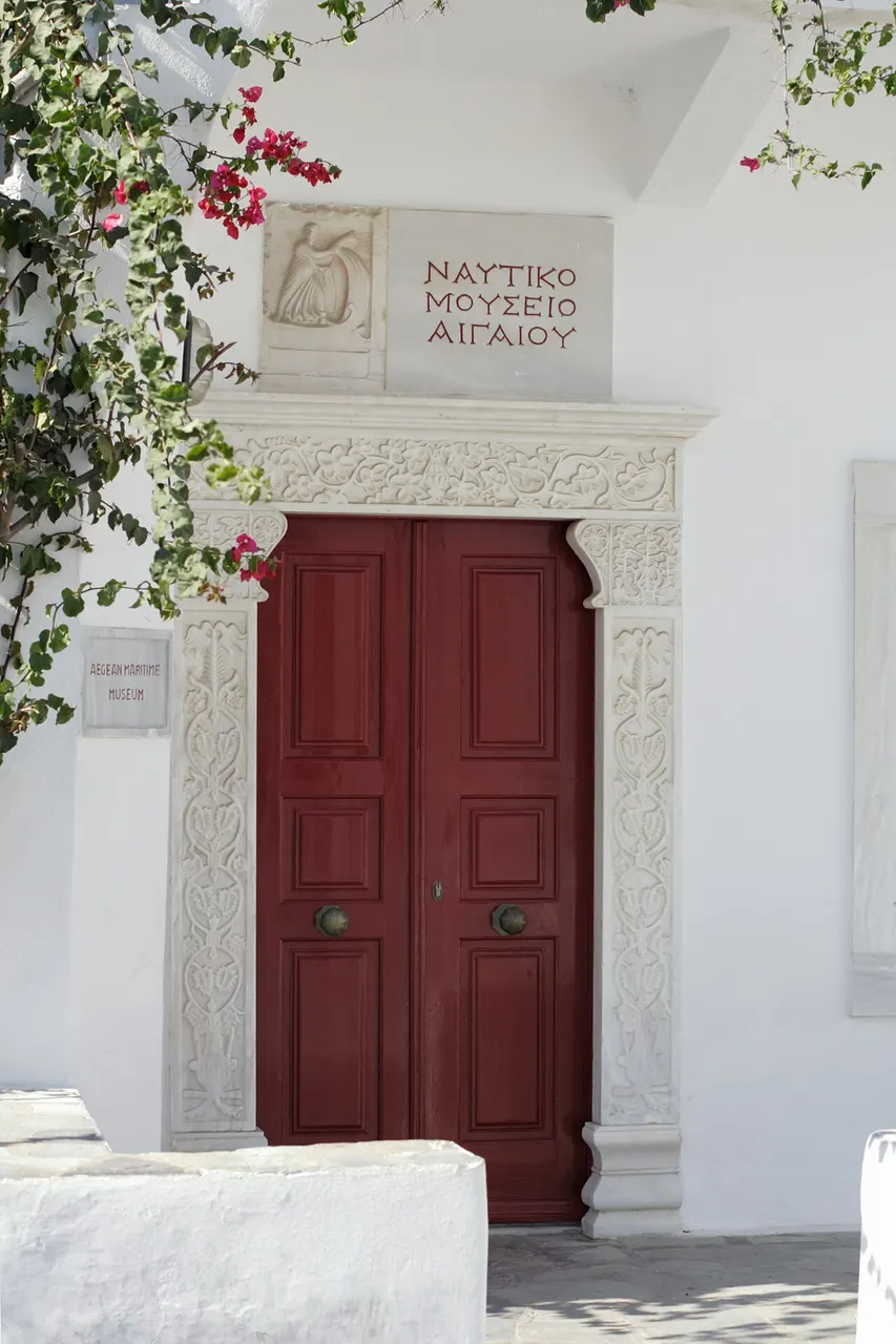 A porta vermelha do Museu Marítimo do Egeu, em Mykonos, numa moldura de pedra clara, com a placa do museu em grego acima e uma buganvília ao lado.