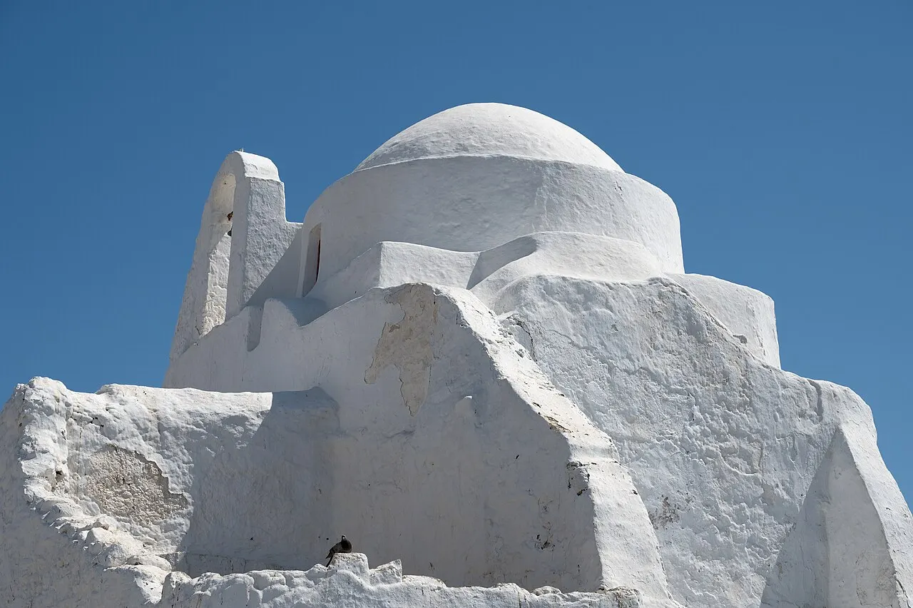 A igreja de Panagia Paraportiani, em Mykonos, um conjunto de capelas caiadas de branco com formas arredondadas e uma cúpula, contra o céu azul.