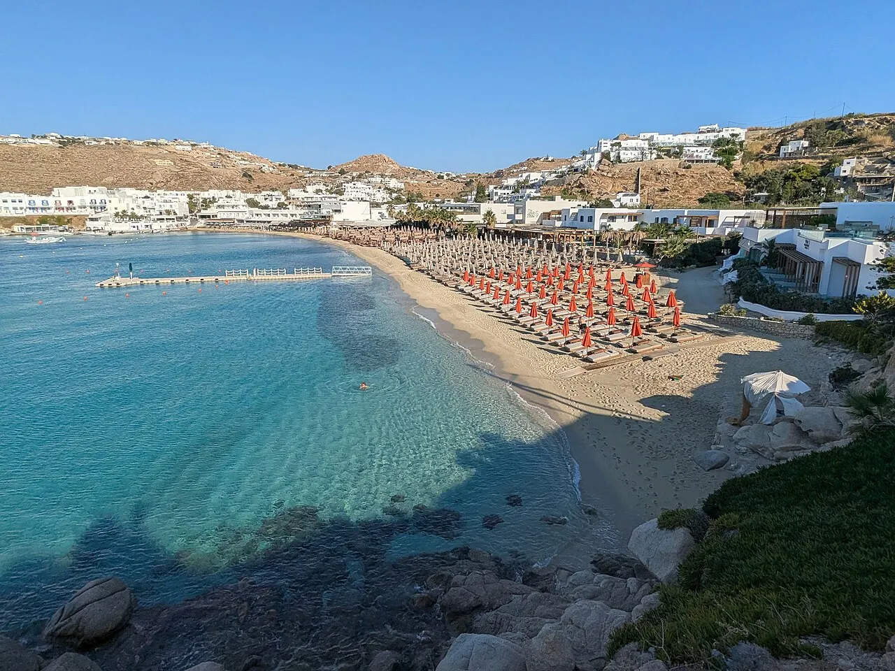 A praia de Platys Gialos vista de cima, com água turquesa transparente, fileiras de guarda-sóis laranja na areia e casas brancas na encosta.