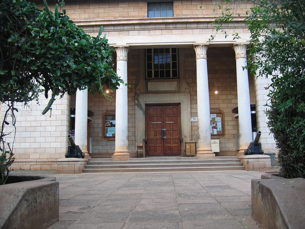 Entrada do Museu Nacional do Quênia: pórtico de quatro colunas coríntias claras e porta dupla de madeira no alto de uma escadaria, com dois canhões antigos nos lados e vegetação densa enquadrando a fachada.