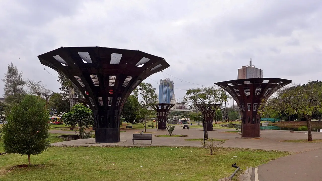Gramado do Uhuru Park, com dois abrigos escuros em forma de funil, bancos e torres da cidade ao fundo.
