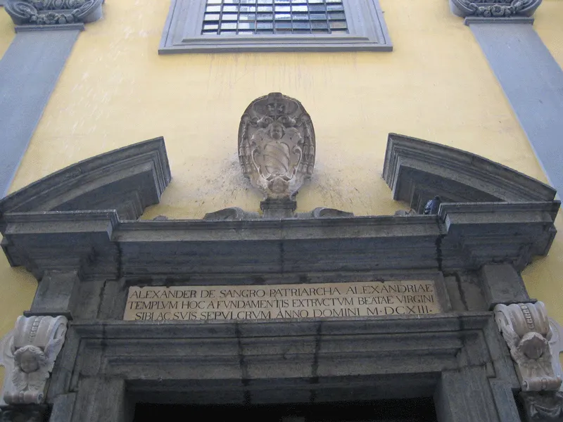 Portal de pedra da Cappella Sansevero, com inscrição latina sobre a porta, um brasão e a parede amarela ao redor.