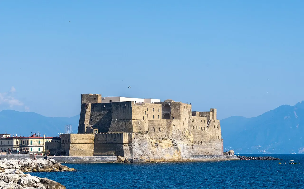 Castel dell'Ovo, em Nápoles, fortaleza de pedra sobre um ilhéu no golfo, com montanhas ao fundo.