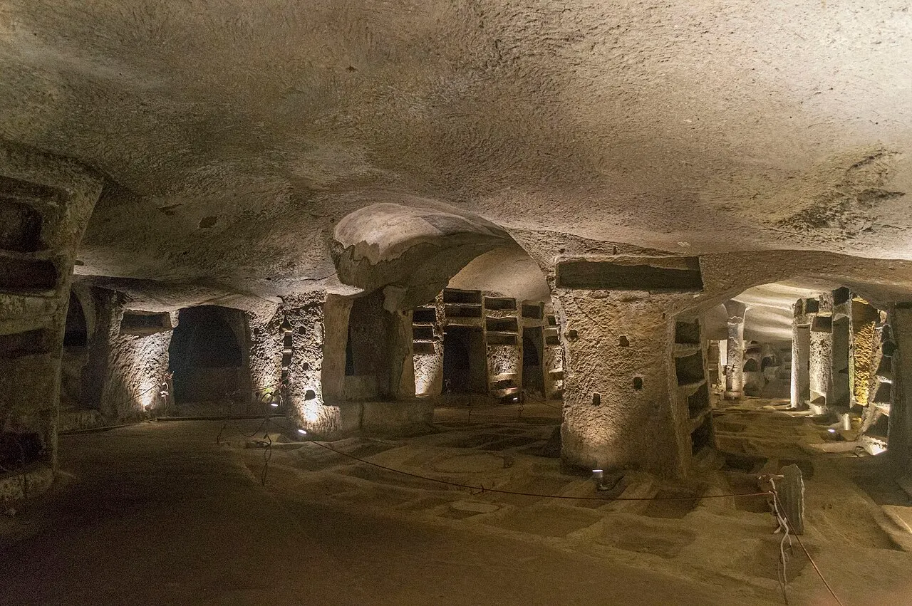 Salão subterrâneo das Catacumbas de San Gennaro, com pilares e nichos escavados na rocha sob luz amarela.