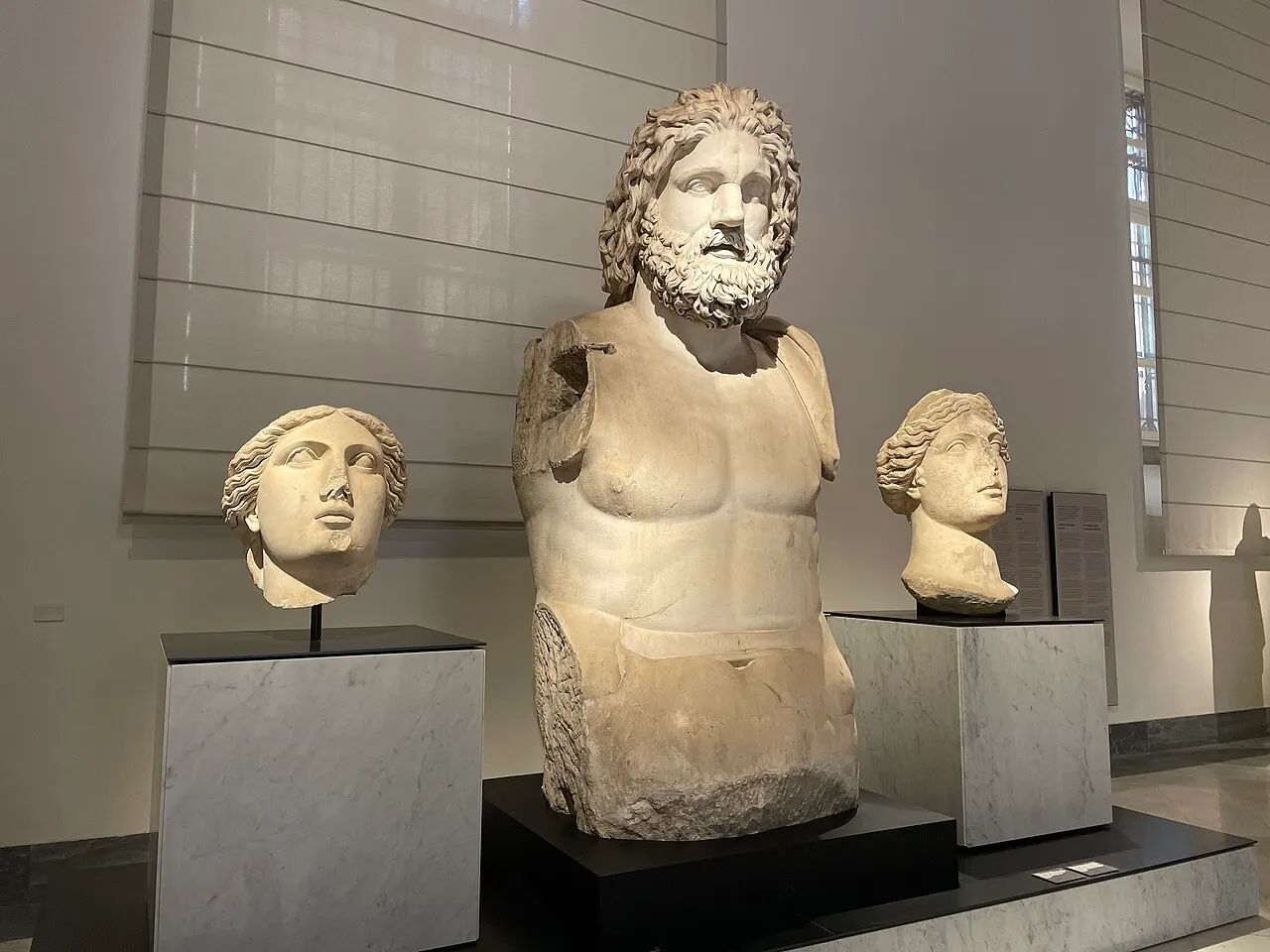 Torso colossal de mármore de um homem barbado sobre base escura, entre duas cabeças antigas em pedestais de mármore, numa sala clara do Museu Arqueológico de Nápoles.