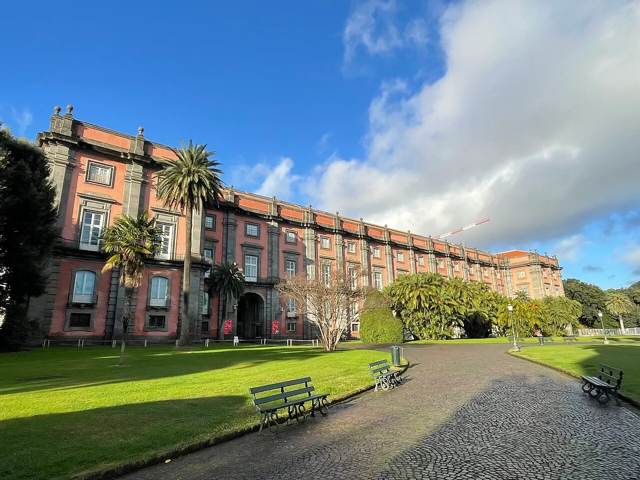 Palácio de Capodimonte, em Nápoles, com a fachada vermelha e cinza em perspectiva, entre palmeiras e gramado.