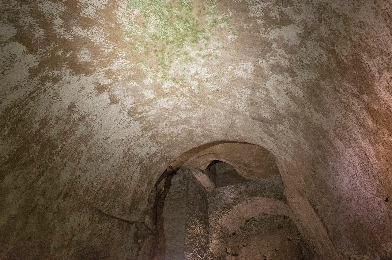 Galeria escavada na rocha de tufo sob Nápoles, iluminada, com uma abertura em arco ao fundo.