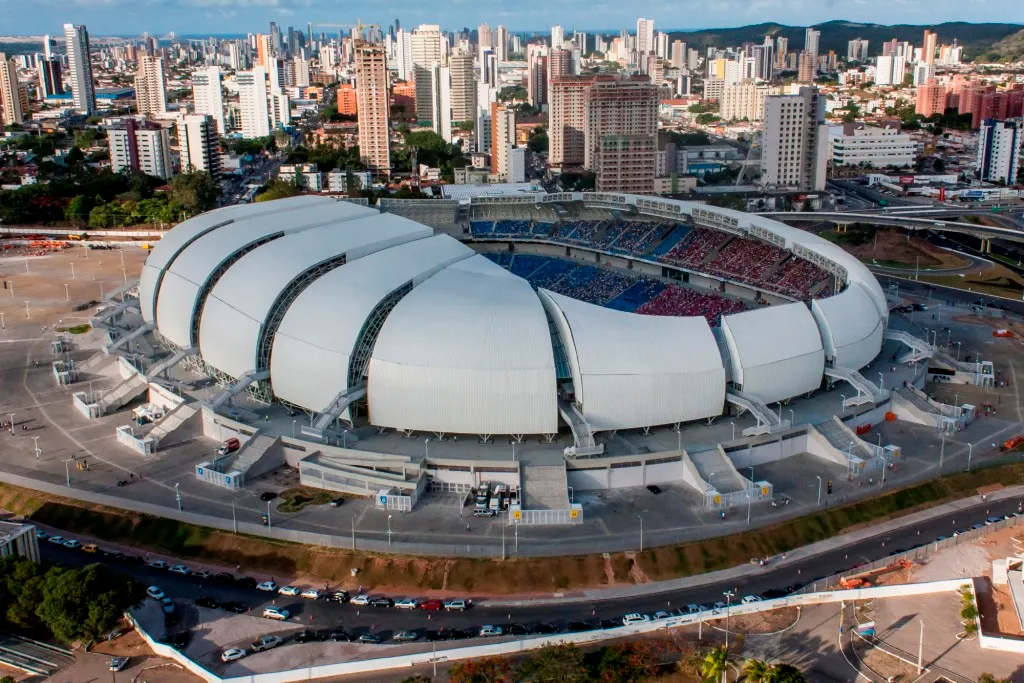 A Arena das Dunas vista do alto, com a cobertura branca em gomos e os prédios de Natal ao fundo.