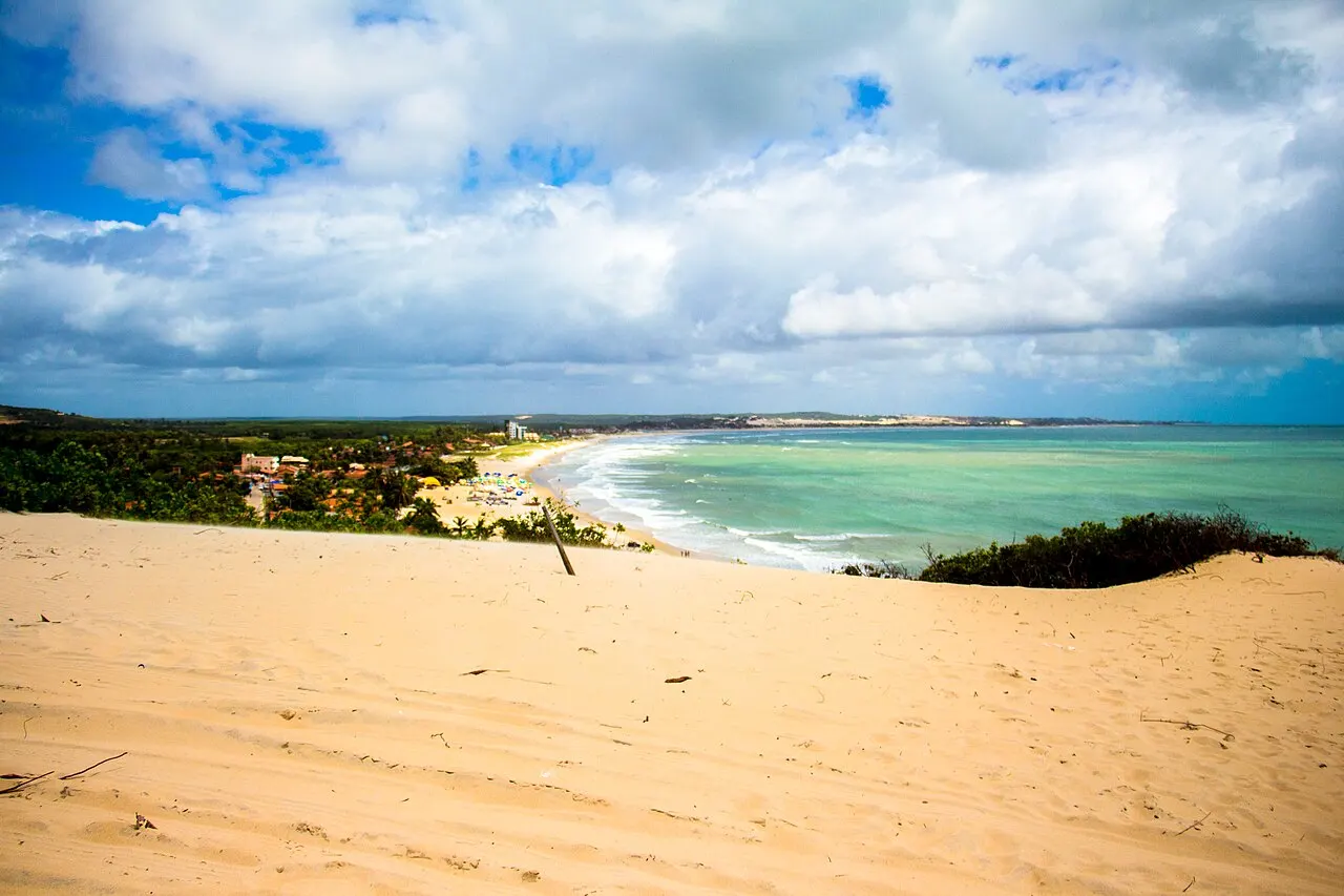 O alto das dunas de Genipabu, de areia clara, com a curva da praia e o mar verde ao fundo sob nuvens.