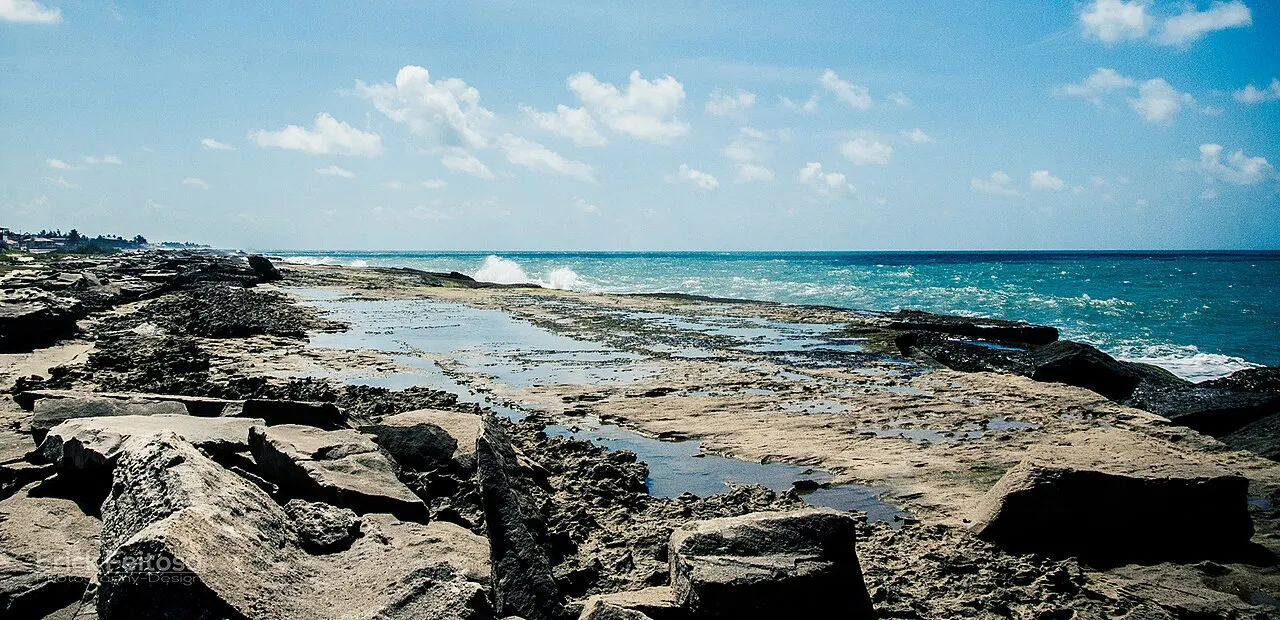 As pedras escuras da beira da Praia de Camurupim, com poças entre elas e as ondas quebrando no mar azul.
