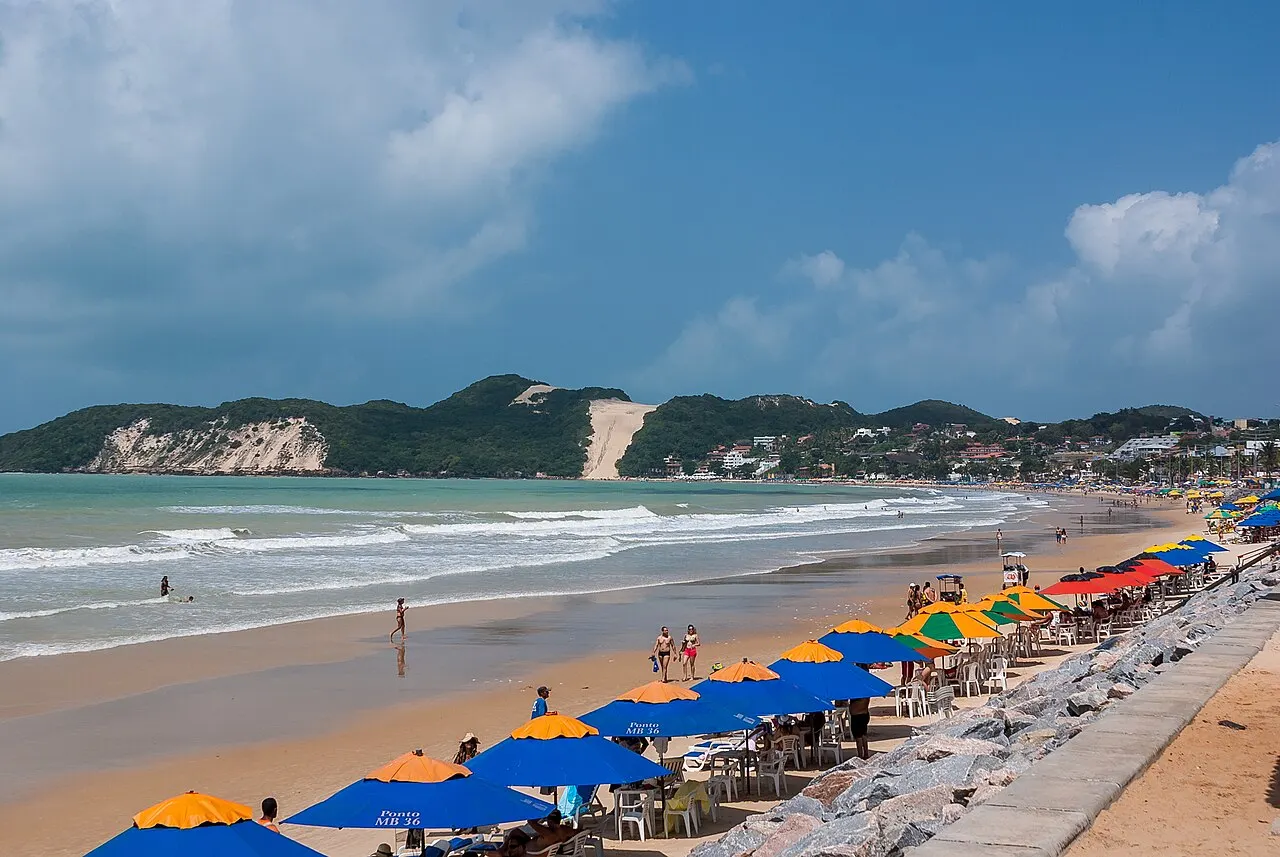 A Praia de Ponta Negra com fileiras de guarda-sóis coloridos na areia e o Morro do Careca, a duna de areia clara, ao fundo.