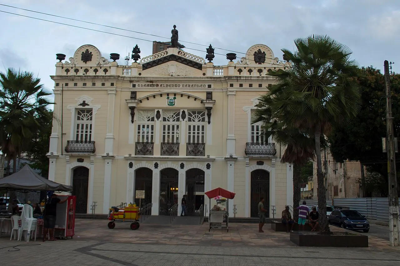 A fachada creme e ornamentada do Teatro Alberto Maranhão, entre palmeiras, na praça em frente.