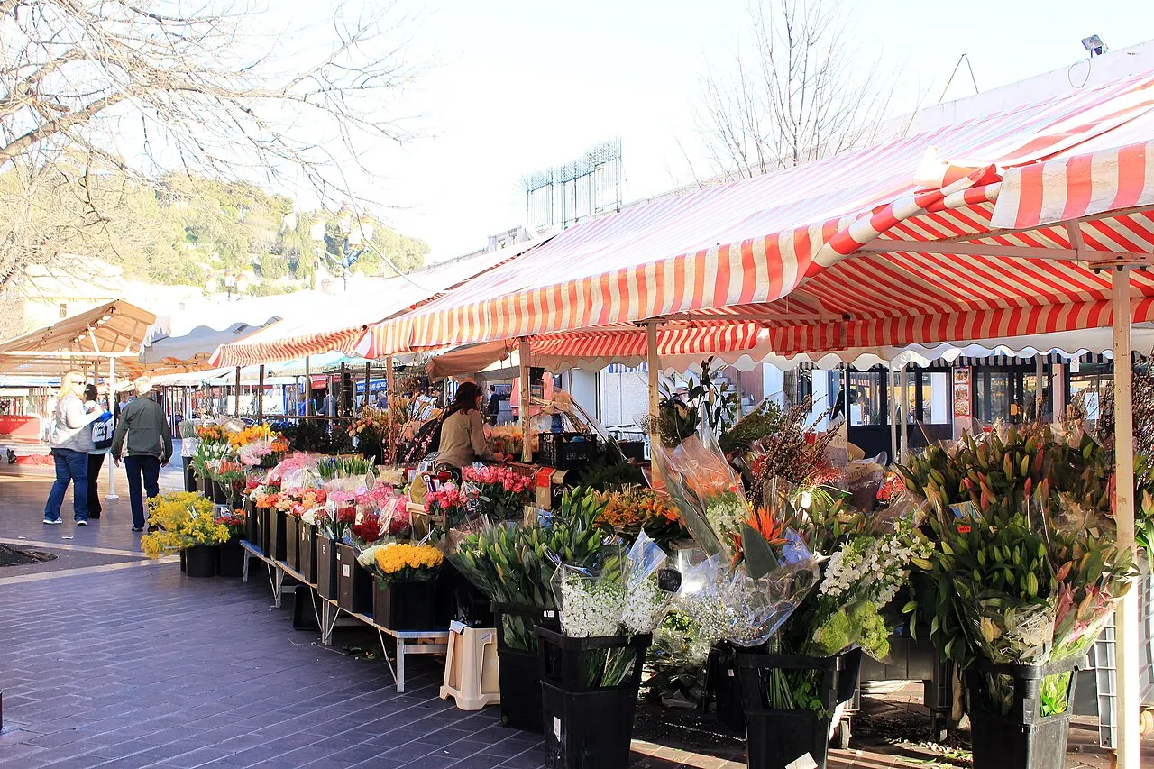 Bancas de flores do Cours Saleya, em Nice, com buquês coloridos em baldes sob toldos listrados de vermelho e branco e pessoas passando entre as barracas.