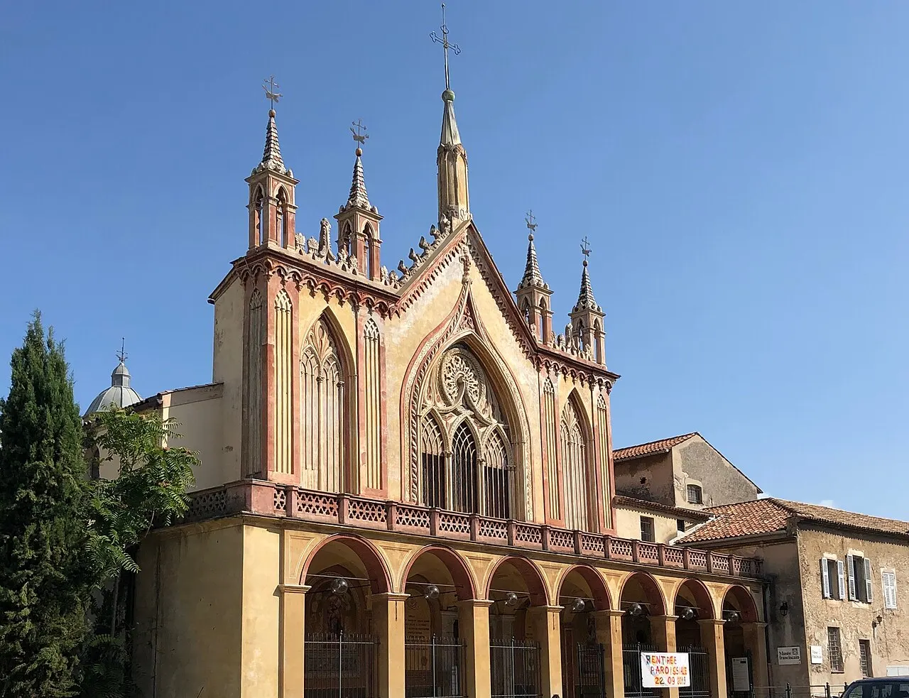 A fachada neogótica da igreja do mosteiro de Cimiez, com pináculos e uma rosácea no alto e um pórtico de arcos na base, sob céu azul.