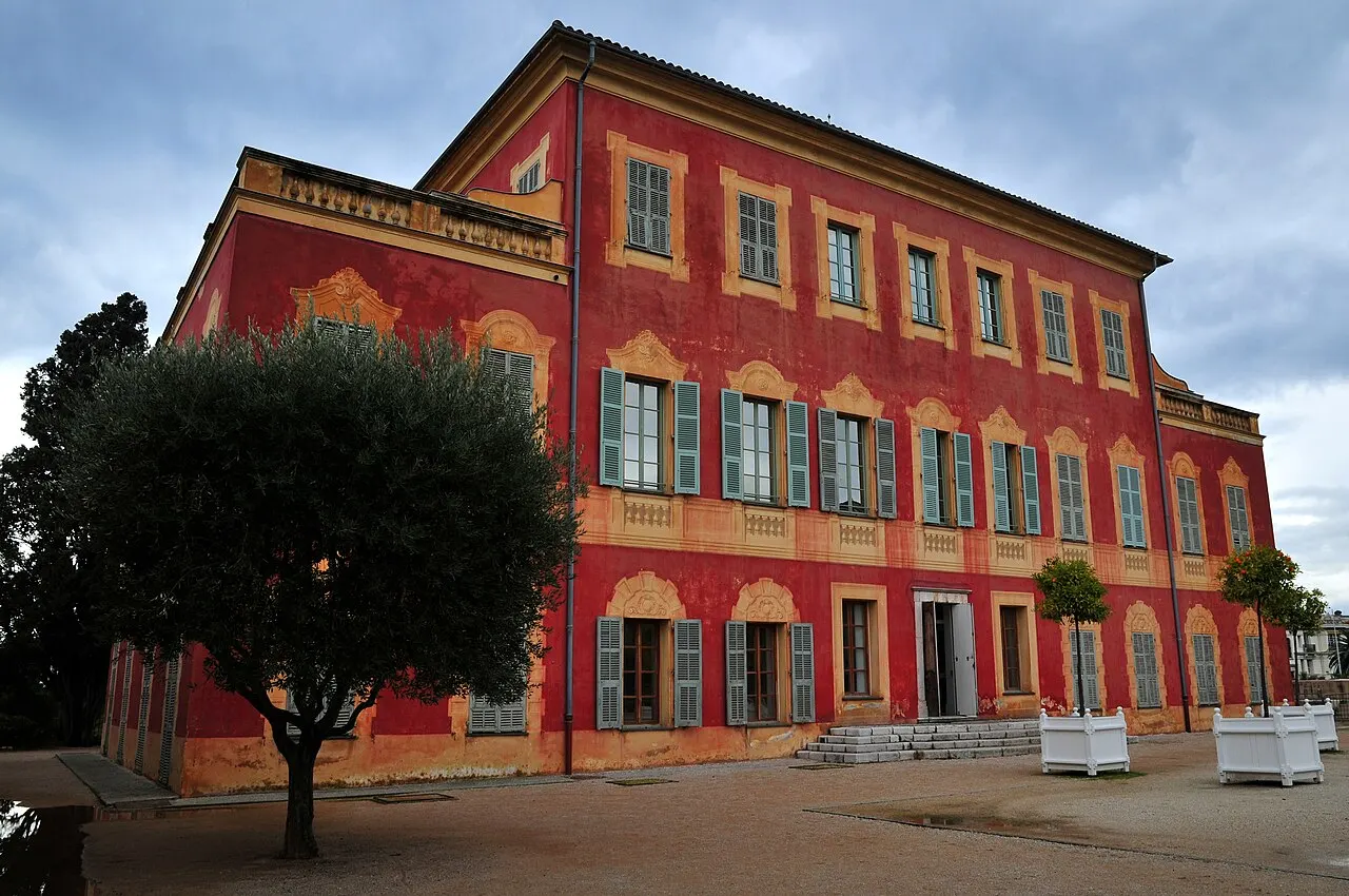 A fachada vermelha da villa genovesa do Museu Matisse, em Cimiez, com janelas de venezianas verdes e molduras amarelas pintadas, e uma oliveira em primeiro plano.