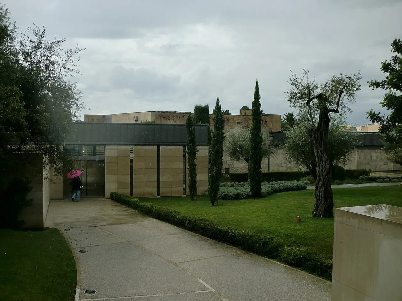 O caminho de entrada do Museu Nacional Marc Chagall, em Nice, entre gramados e oliveiras, com o prédio baixo de pedra clara ao fundo, sob céu nublado.