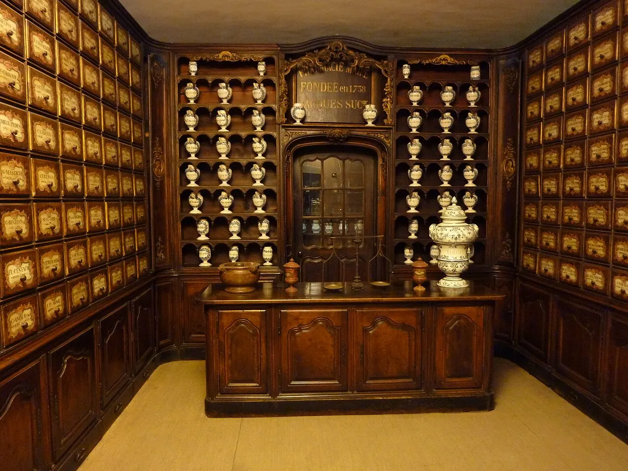 Uma farmácia antiga remontada no Palais Lascaris, com estantes de madeira escura, gavetas etiquetadas e potes de cerâmica brancos em prateleiras atrás do balcão.