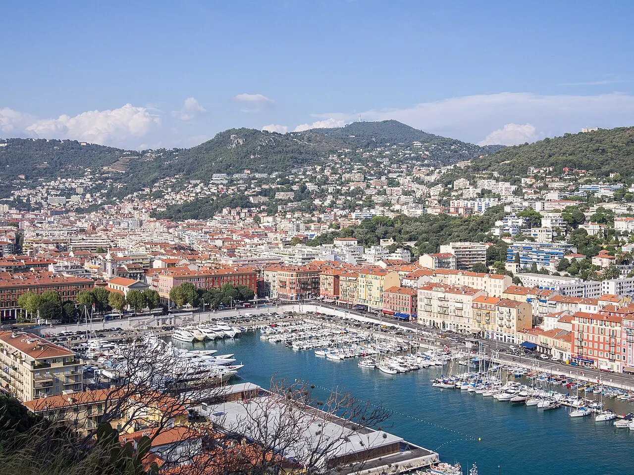 O Port Lympia, em Nice, visto do alto, com barcos atracados na água azul, prédios de fachadas ocre e rosadas em volta e colinas cobertas de casas ao fundo.