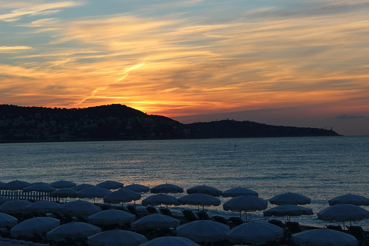 A Baie des Anges ao amanhecer, com céu alaranjado, silhueta da costa ao fundo e guarda-sóis no primeiro plano, vista da Promenade des Anglais.