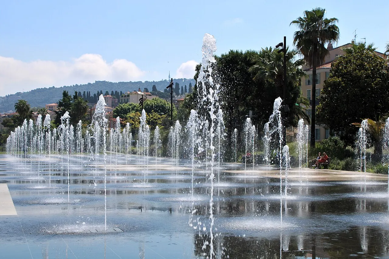 Os jatos d'água do espelho d'água da Promenade du Paillon, em Nice, refletindo o céu, com palmeiras e prédios ao fundo.