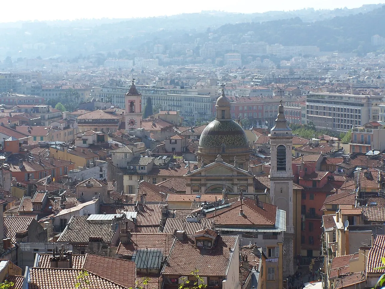 Os telhados de telha de Vieux Nice vistos do alto, com uma cúpula, campanários e prédios mais novos e colinas ao fundo, sob uma névoa clara.