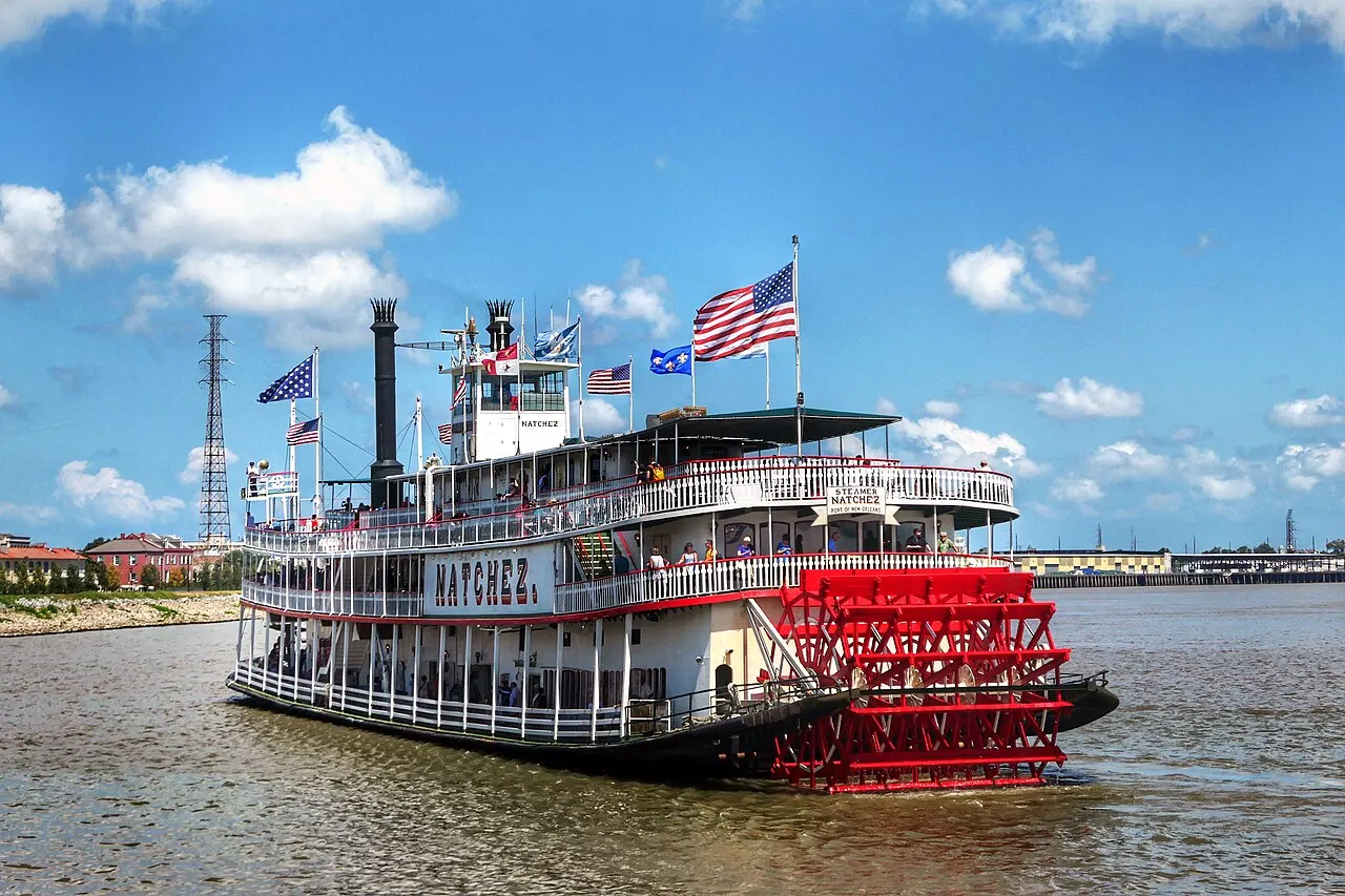 Vapor de rodas vermelho e branco navega o rio Mississippi, com bandeiras dos Estados Unidos e da Louisiana hasteadas no convés superior sob céu azul.