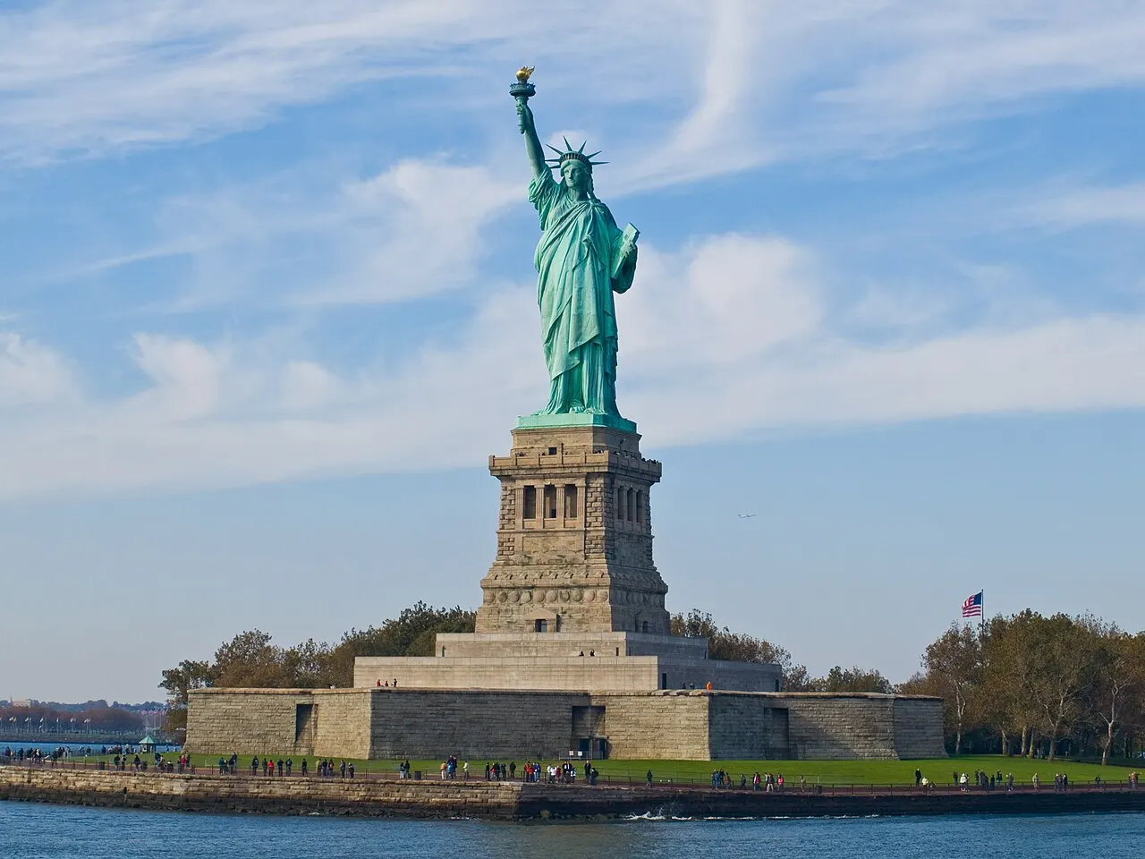 Estátua da Liberdade, em Nova York, verde sobre o pedestal de pedra, na ilha, com a água em volta.