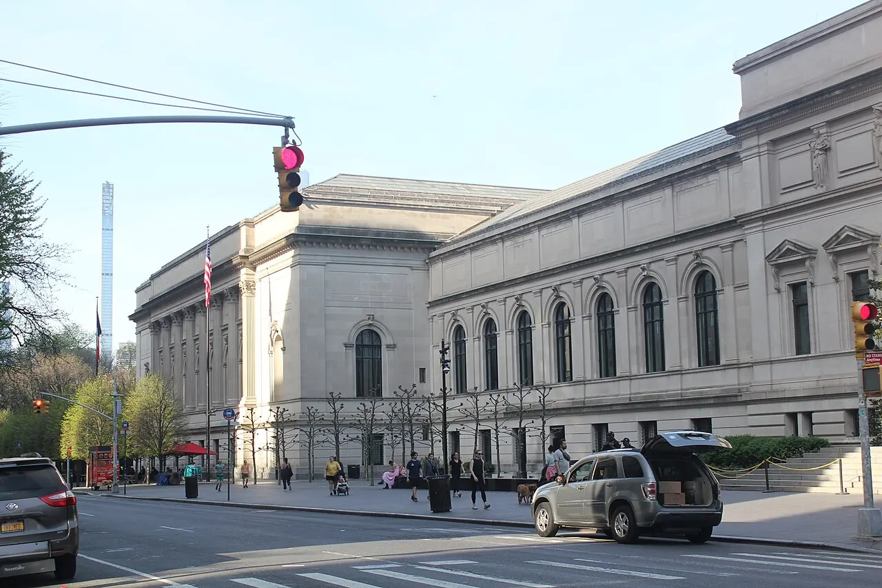 Fachada de pedra clara do Metropolitan Museum of Art, com janelas em arco, uma bandeira hasteada e pessoas caminhando na calçada ao entardecer.