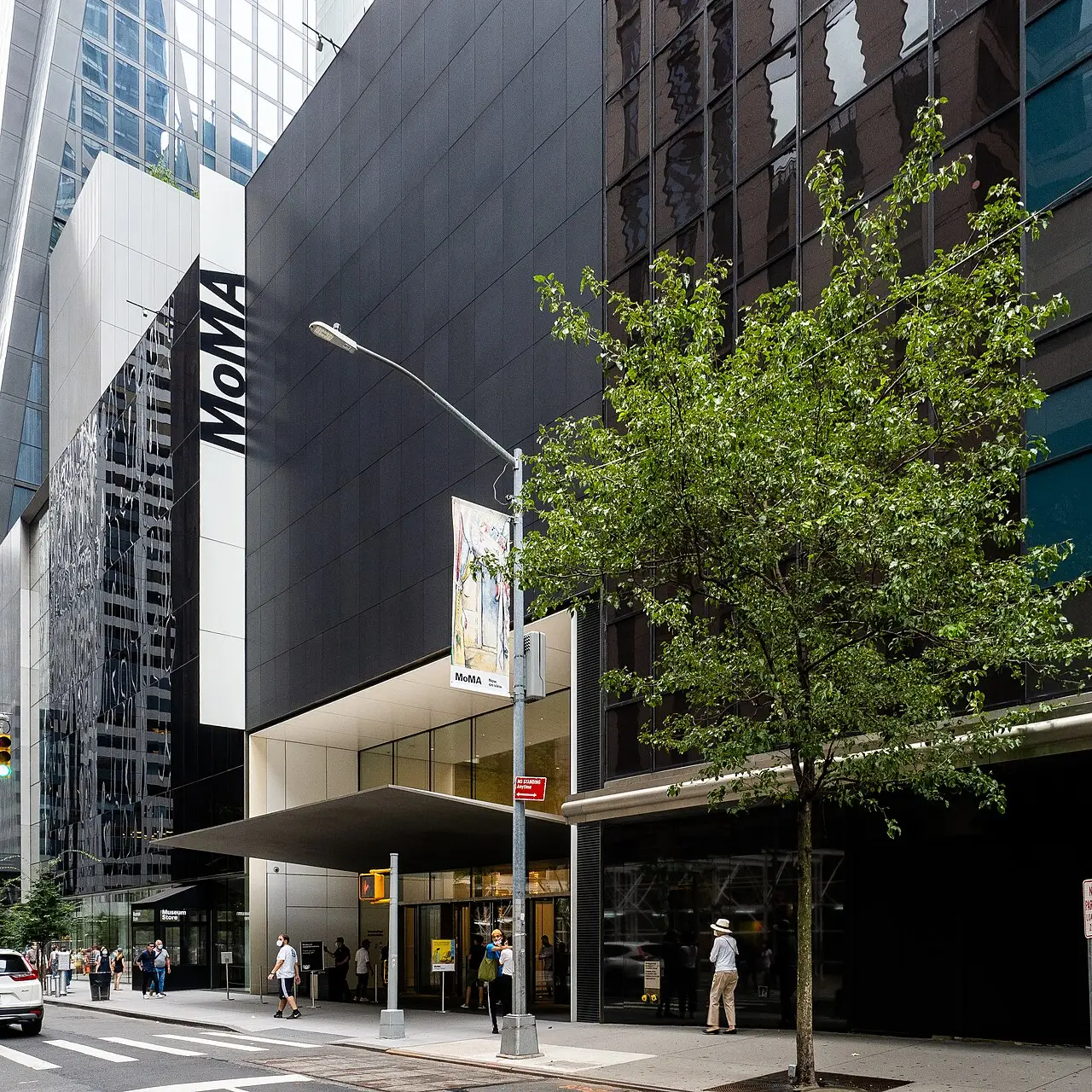Fachada do MoMA, em vidro e painéis escuros, entre edifícios altos, com uma árvore na calçada.