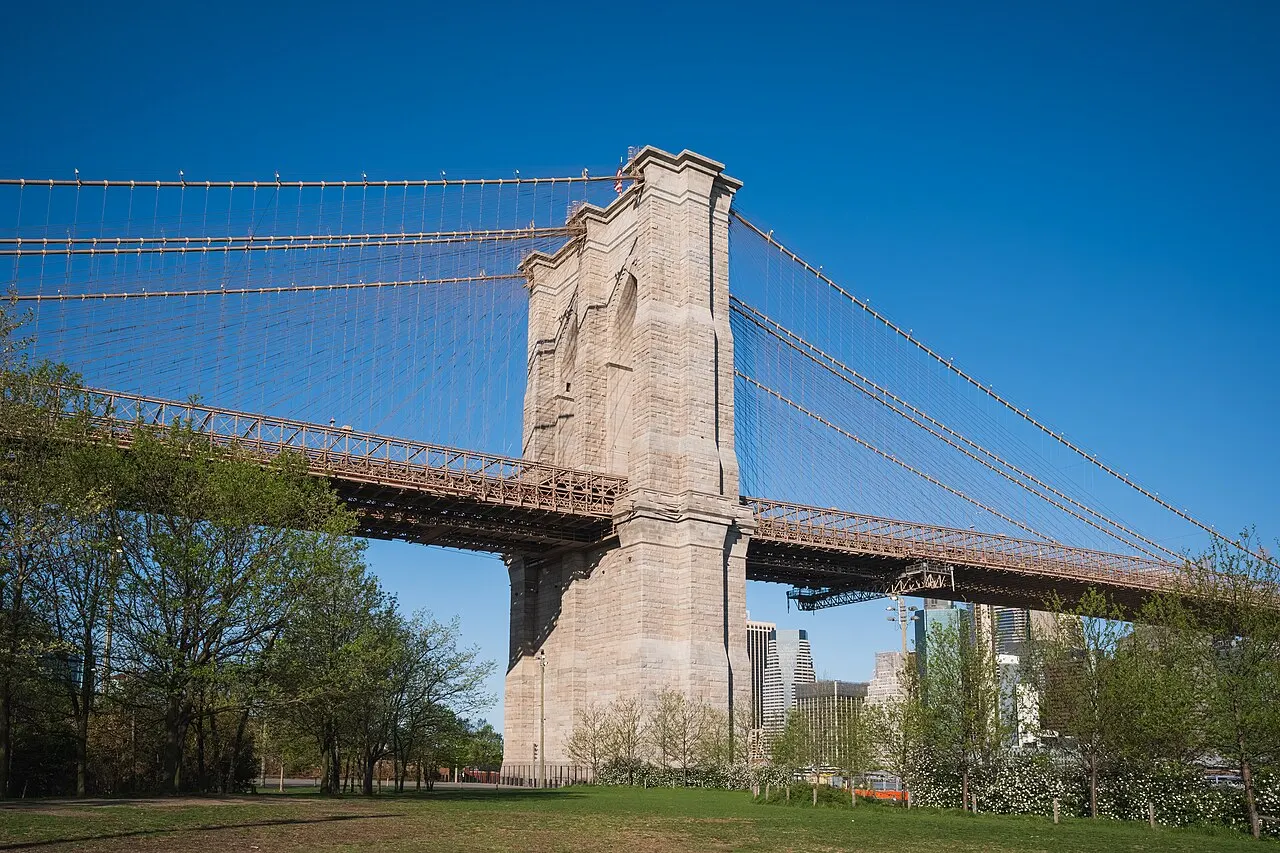 Torre de pedra da Ponte do Brooklyn com os cabos de suspensão abertos em leque, árvores em primeiro plano e arranha-céus ao fundo sob céu azul sem nuvens.