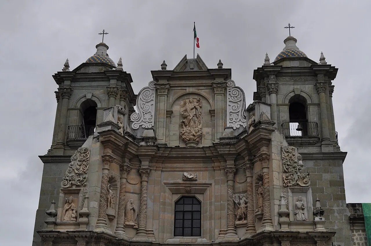 Fachada barroca de pedra verde da basílica de Nuestra Señora de la Soledad, em Oaxaca, com duas torres de sino de cúpula azul e amarela, santos em nichos e um relevo da Virgem no alto, sob céu encoberto.