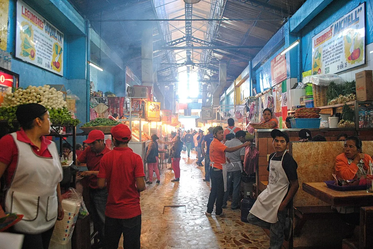Corredor coberto de mercado com fumaça de churrasco no ar, vendedores usando aventais atendendo clientes entre bancadas de carne grelhada e barracas iluminadas.