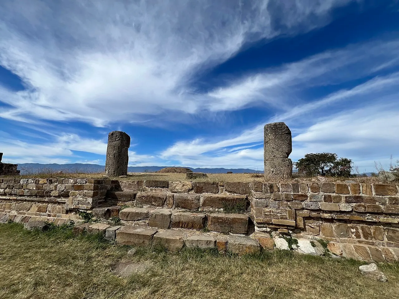 Muros baixos de pedra e duas colunas cilíndricas de Monte Albán em primeiro plano, com a planície e a serra de Oaxaca ao fundo sob céu azul e nuvens finas.