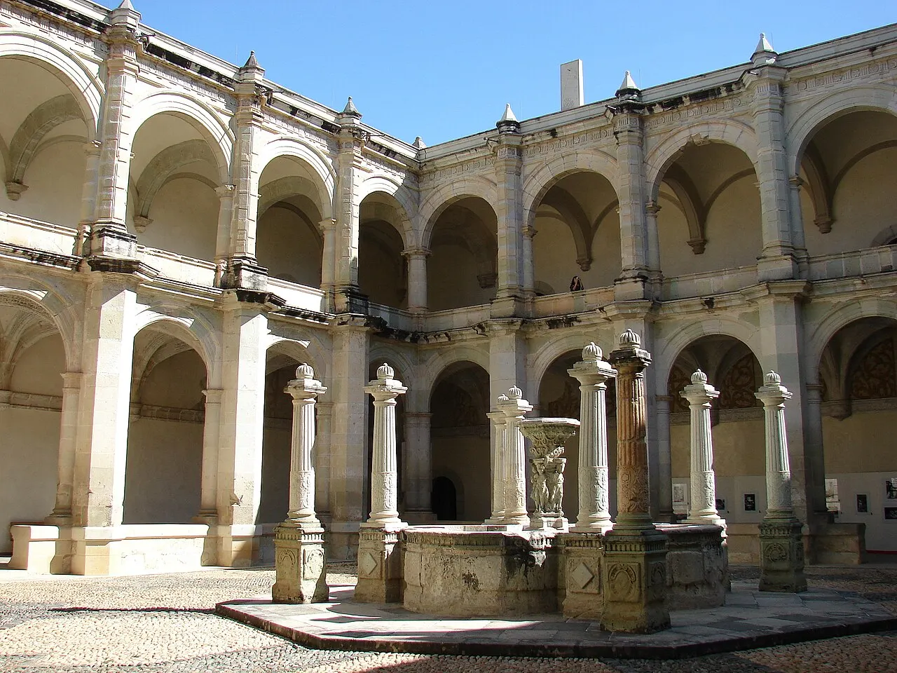 Pátio de dois andares com galerias de arcos de pedra clara do antigo convento de Santo Domingo, com uma fonte esculpida ao centro sob céu azul.
