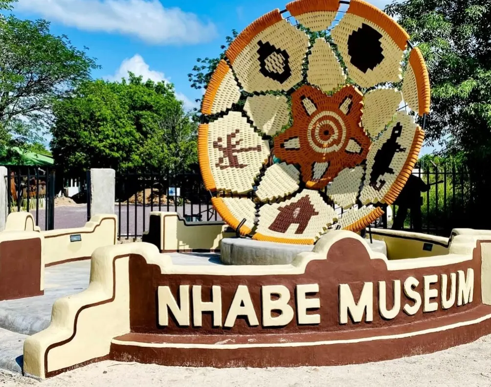 Large sculpture shaped like a woven basket, in cream, orange and brown with animal and star motifs, on a base with the letters "NHABE MUSEUM", in front of a railing gate and trees.