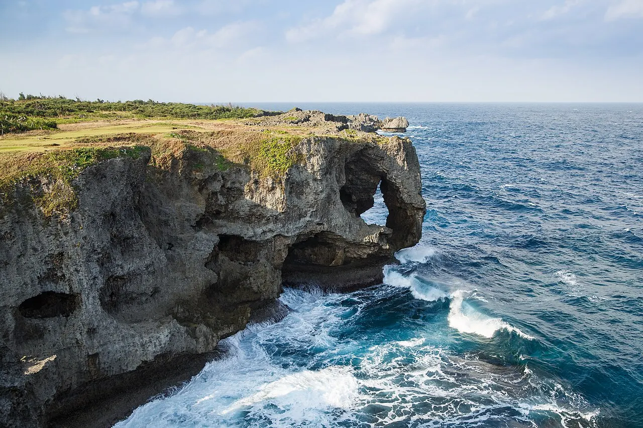 The limestone cliff of Cape Manzamo, with an arch opened in the rock and a grass-covered top, above the blue sea with waves breaking at its base.