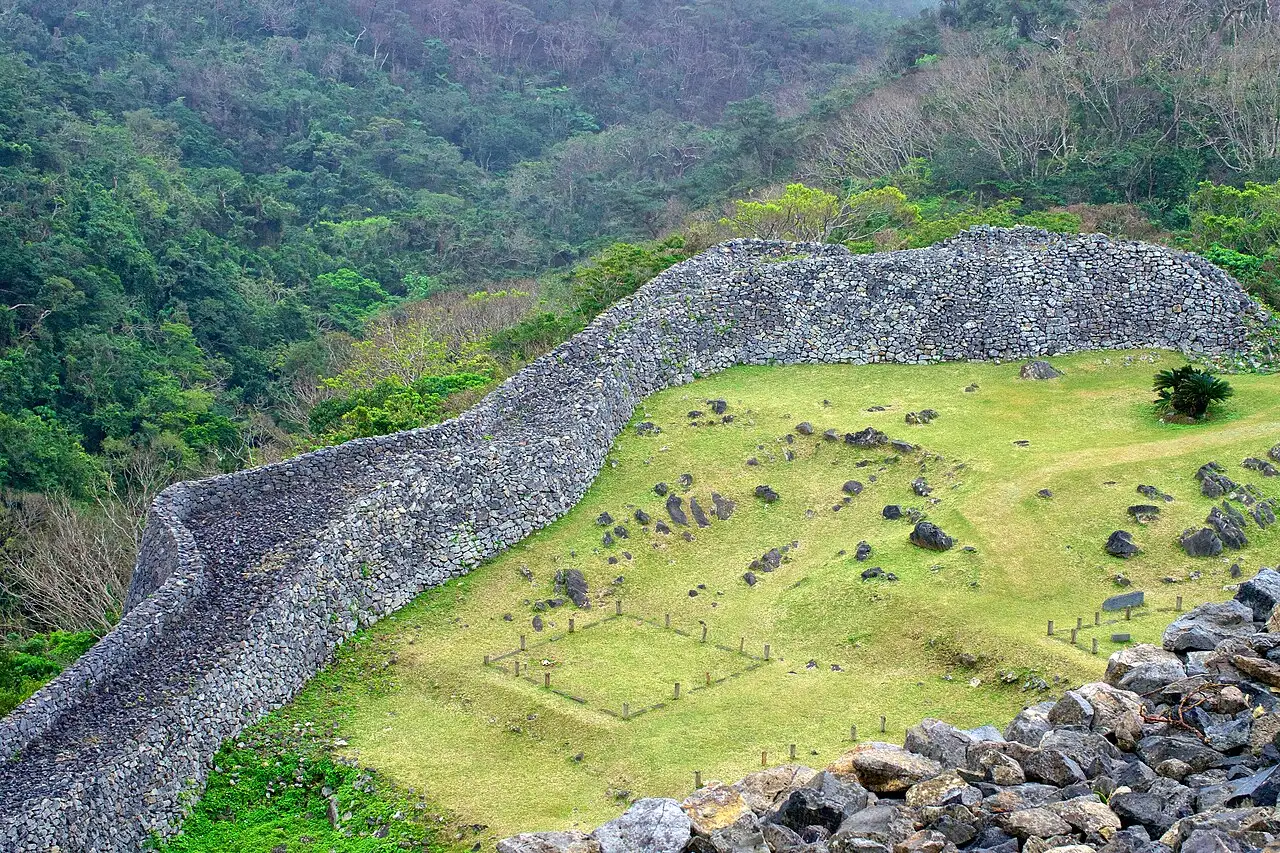 The stone wall of Nakijin Castle winding along the slope, enclosing a grassy area with loose stones, with forest behind.